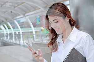 Happy young Asian business woman using mobile smart phone at sidewalk of office.