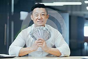 A happy young Asian businessman holds a cash of money in his hands and waves it, sitting in a office