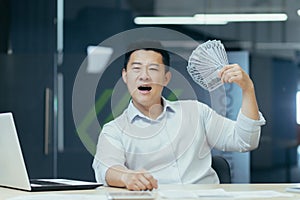 A happy young Asian businessman holds a cash of money in his hands and waves it, sitting in a office