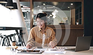 Happy young asian businesswoman sitting on her workplace and using calculator in the office. Young woman working at laptop in the