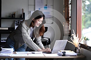 Happy young Asian businesswoman standing using calculator and laptop computer at office.