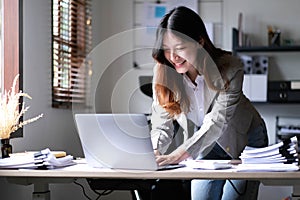 Happy young Asian businesswoman standing using calculator and laptop computer at office.