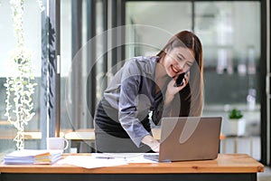Happy young Asian businesswoman standing using smartphone and laptop computer at office.