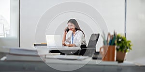 Happy young asian businesswoman talk on the mobile phone and smile while sitting at working place in office