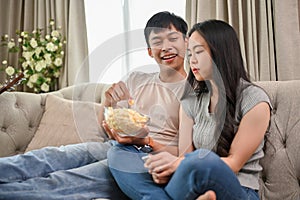 Happy young Asian couple enjoy eating popcorn and watching a movie while relaxing on a sofa