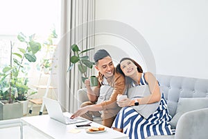 Happy young Asian Couple At Home Using Laptop Computer when sitting on sofa