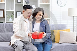 Happy young Asian couple, man and woman sitting on sofa at home. A man gives a woman a red box as a surprise gift. They