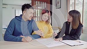Happy young Asian couple and realtor agent. Cheerful young man signing some documents while sitting at desk together with his wife
