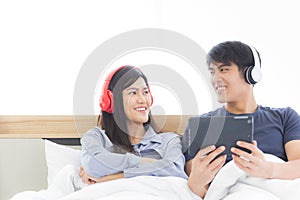 Happy young Asian couple is sitting in bed and enjoying listening to songs or watching movies on a tablet computer in the bedroo