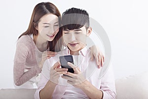 Happy young Asian couple using cellphone, browsing internet, chatting online, relaxing on sofa at home