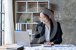 Happy young asian entrepreneur woman in glasses counting profit, on calculator at laptop computer, analyzing benefits