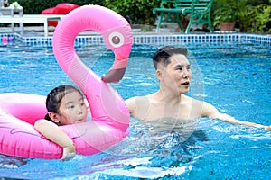Happy young Asian father and little girl daughter with pink flamingo plastic toy in blue water pool, dad teaching child kid