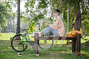 A happy young Asian female college student using her digital tablet on a bench in a green park