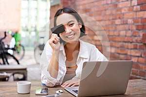 Happy young asian female, sitting with laptop and showing credit card, paying bills online, shopping in internet