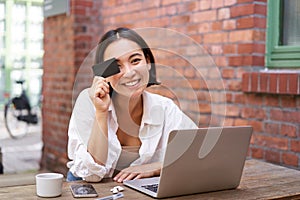 Happy young asian female, sitting with laptop and showing credit card, paying bills online, shopping in internet