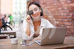 Happy young asian female, sitting with laptop and showing credit card, paying bills online, shopping in internet