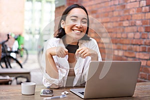 Happy young asian female, sitting with laptop and showing credit card, paying bills online, shopping in internet