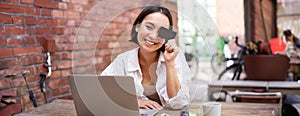 Happy young asian female, sitting with laptop and showing credit card, paying bills online, shopping in internet