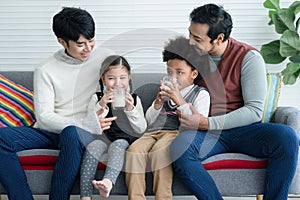 Happy young Asian gay couple with diverse adopted children, African and Caucasian, drinking milk and sitting on sofa at home.