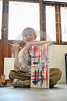 A happy young Asian gay man sitting on the gallery floor with his painted canvas