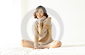 Happy young Asian girl with curly hair wearing knitted warm sweater and looking at camera in her bedroom. Concept woman lifestyle