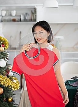 Happy young Asian girl trying to choose a dress standing next to Xmas-tree