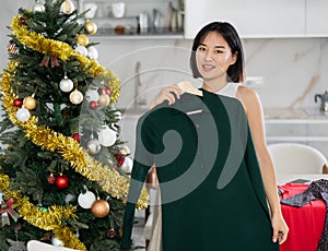 Happy young Asian girl trying to choose a dress standing next to Xmas-tree