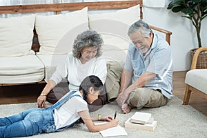 Happy young asian granddaughter reading and writing book with grandfather and grandmother looking beside on floor in living room