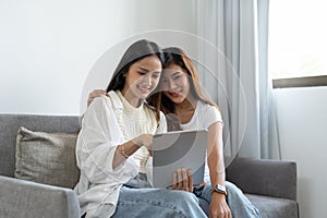 Happy young asian lesbian couple hugging having fun using digital tablet relaxing on couch at home. Two smiling women