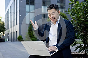 A happy young Asian male businessman is sitting outside an office building and smilingly looking at the screen of a photo