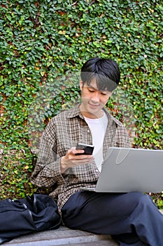 Happy young Asian male college student in the city park, using his smartphone and laptop