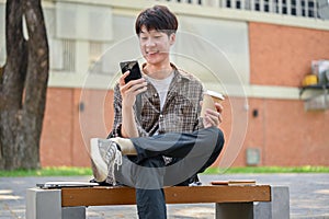 A happy young Asian male college student sits on a bench in the campus park using his smartphone