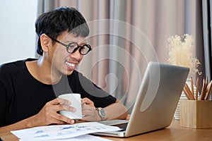 Happy young asian man holding coffee cup and using computer laptop