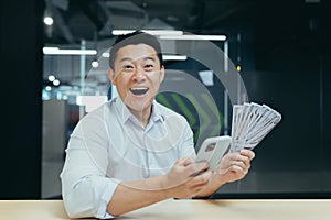A happy young Asian man holds a telephone and cash money in his hands in office
