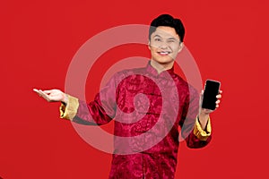 Happy young Asian man in red cheongsam qipao showing mobile phone isolated on red background