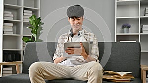 A happy young Asian man using his digital tablet while relaxing on a sofa in the living room