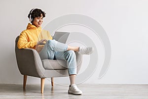 Happy young Asian man using laptop, wearing headphones while sitting in armchair against white wall, copy space