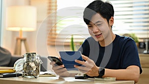 Happy young asian man using smart phone for making online payments, managing expenses finances in living room