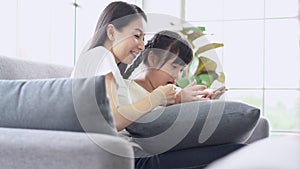 Happy young Asian mother and daughter singing and looking note on mobile while sitting on sofa in living room.
