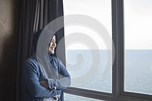 Happy young Asian muslim woman smiling while facing large glass window looking at peaceful ocean view