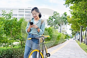 Happy young Asian office woman while riding a bicycle in a city park. She smiles and uses her smartphone