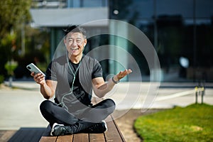 A happy young Asian sportsman man couch is sitting resting on a wooden bench near the lake and using a mobile phone