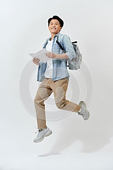 Happy young Asian student man jumping in air with backpack and holding book over white background