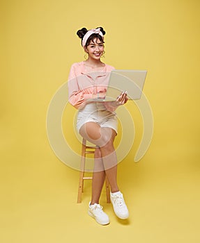 Happy young Asian teen woman smiling. While her using laptop sitting on white chair isolate on bright yellow background