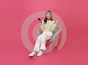 Happy young Asian teen woman smiling listening music in headphones and sitting on chair isolate on pink background