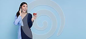 Happy young Asian teen woman talking mobile phone and showing credit card isolated on studio blue background