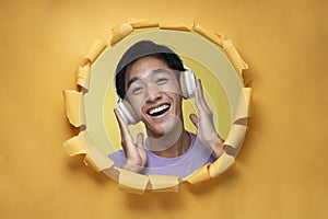 Happy young Asian teenager man laughing poses through torn yellow paper hole with a copy space, wearing purple t-shirt and