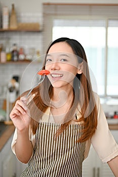 Happy young Asian woman in apron holding a fork with tomato slide, enjoys eating in the kitchen