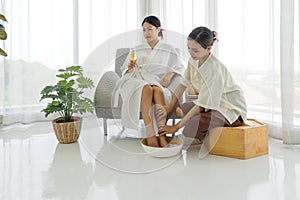 Happy young asian woman in bathrobe with glass of champagne having fun with foot bath from cosmetologist during procedure in spa