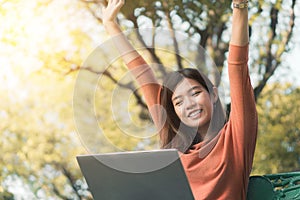 Happy young asian woman be smile working with her laptop and hand up on a bench in the park outdoors on vacation time